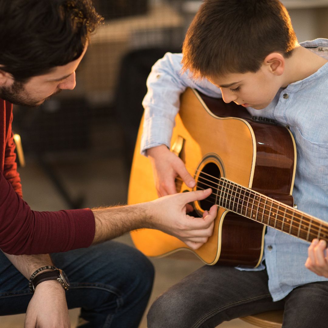 Ein Gitarrenlehrer unterstützt ein Kind beim Gitarre lernen.