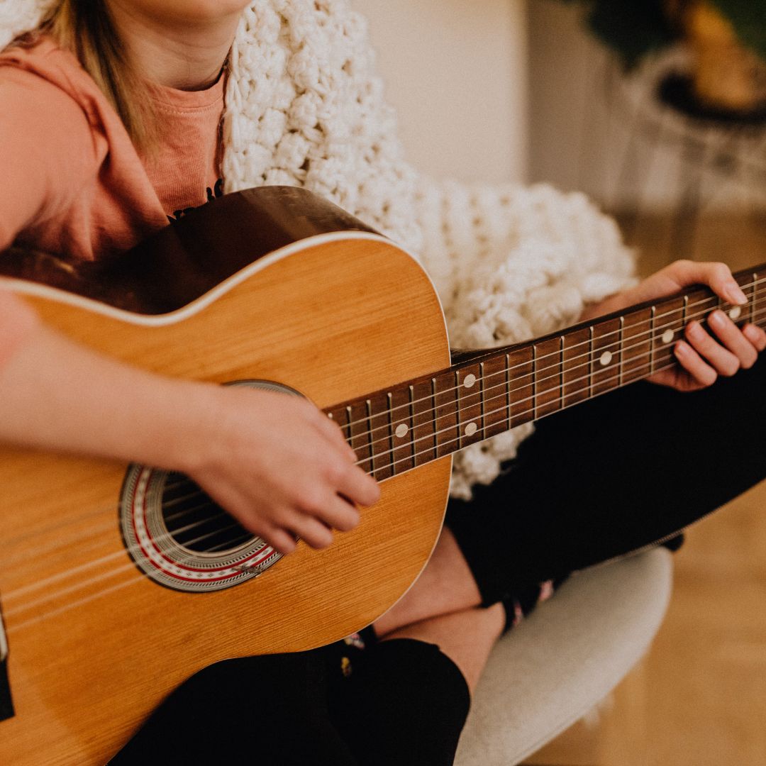 Ein Mädchen, welches im Schneidersitz auf einem Sessel sitzt und eine Akustikgitarre in der Hand hält.
